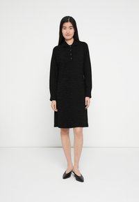 Robe noire à manches longues avec un motif texturé, col polo et trois boutons. La robe arrive juste au-dessus des genoux, associée à des chaussures noires.