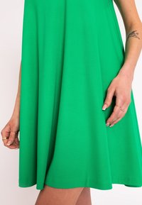Grünes ärmelloses Kleid aus weichem Stoff, mit fließender A-Linie und glatter Textur, während eine Hand am Saum ruht.