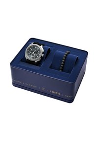 Montre Fossil présentée dans une boîte en métal bleue. Comprend une montre chronographe en ton argent avec un bracelet en caoutchouc noir et un bracelet en perles noires.