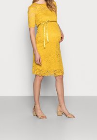 Gul spetsklänning med korta ärmar, vågig nederkant och matchande bälte. Tillsammans med beigh blockklacksandaler för att skapa en enhetlig look.