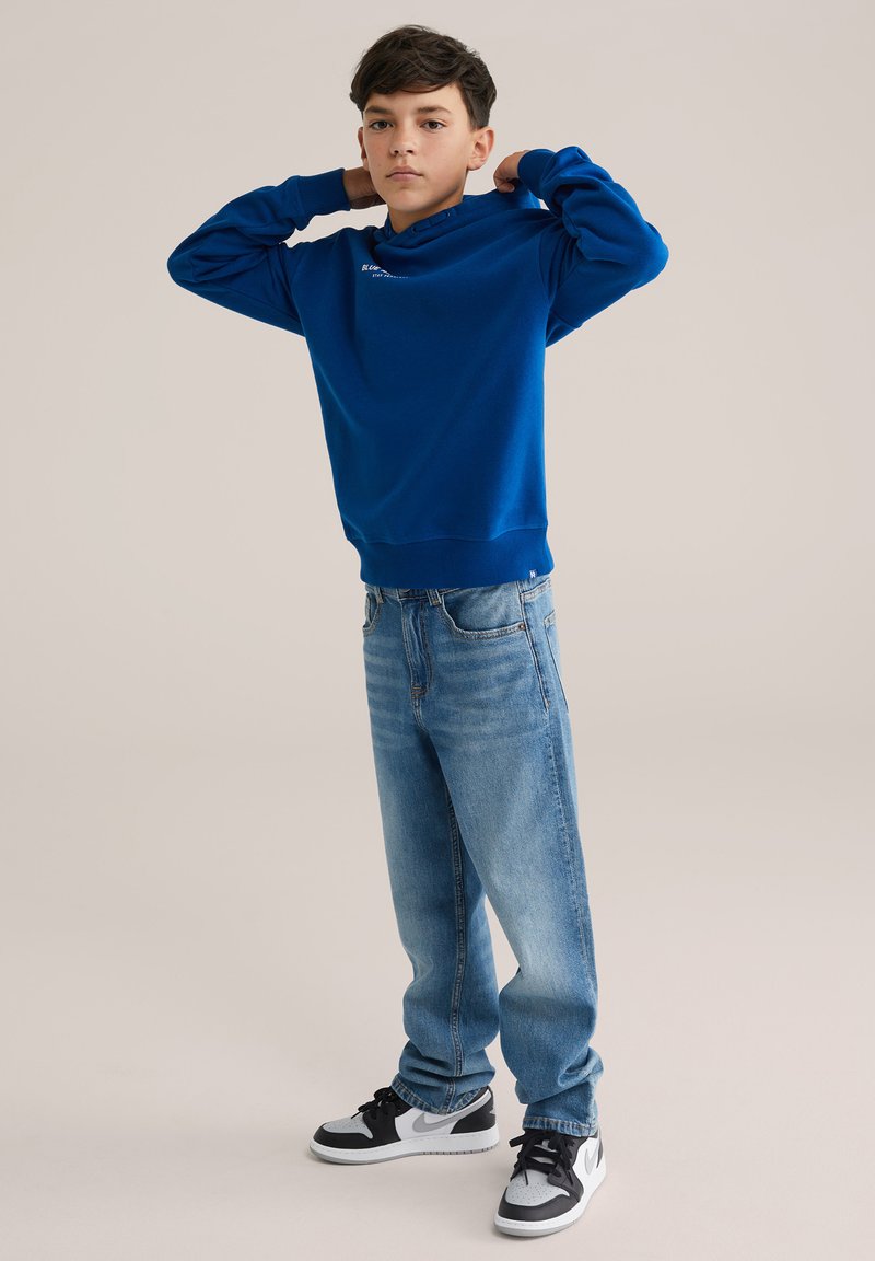 Kék kapucnis pulóver elülső kenguruzsebbel és bordázott mandzsettákkal, világoskék, laza szabású farmernadrággal és fekete-fehér sportcipőkkel párosítva.