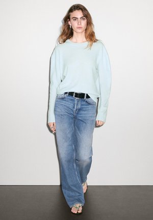 Hellblaues Oversize-Pullover, hoch taillierte blaue Jeans, schwarzer Gürtel und grüne Flip-Flops. Das Modell steht vor einem einfarbigen Hintergrund.