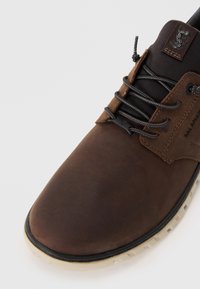 Chaussure en cuir marron avec un bout arrondi, langue en tissu noir et lacets gris. Elle possède une semelle en caoutchouc blanche et un détail de logo en relief.