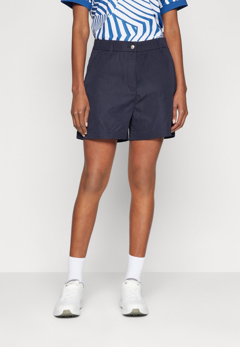 Pantaloni corti blu navy con una vestibilità sartoriale, dotati di chiusura con bottone frontale, passanti per cintura, tasche laterali e orli arrotolati. Indossati con una camicia blu a fantasia e sneakers bianche.