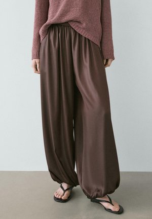 Kobieta nosząca luźne brązowe jedwabne spodnie z elastycznymi nogawkami, różowy dziany sweter i czarne sandały typu thong, stojąca na szarej podłodze.