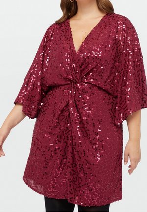 Vrouw met een dieprode jurk met pailletten, driekwart mouwen en een geploochte knoop in de taille, waarbij het bovenlichaam en de armen zichtbaar zijn.