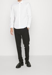 Chemise blanche boutonnée avec col, associée à un pantalon noir ajusté et des mocassins noirs. Design simple sans motifs ni textures visibles.