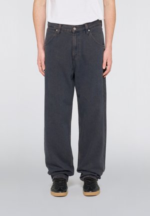 Personne portant un jean noir ample et des baskets noires avec des semelles en gomme, debout devant un fond blanc uni.