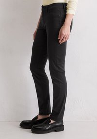 Zwarte slim-fit jeans met een gladde textuur, voorzien van zijzakken en gestikte naden, gecombineerd met zwarte instapschoenen op een lichte achtergrond.