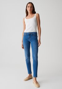 Jean en denim bleu skinny avec une taille mi-haute, un léger délavage, un design à cinq poches et des effets de fausse usure. Associé à un débardeur blanc et des ballerines beiges.