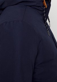 Mörkblå jacka med en slät yta, elastiskt midjeband och kontrasterande dragsnöre med metalländar. Fickan är synlig på sidan.