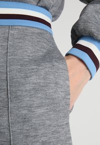 Un pantalon de survêtement gris présente une taille côtelée avec des rayures bleues, blanches et bordeaux. Le tissu a un aspect texturé et comprend des poches latérales.