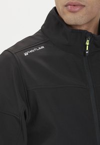 Czarna kurtka typu softshell z wysokim kołnierzem, z przednim zamkiem błyskawicznym i odblaskowym logo na lewym ramieniu. Trwała, gładka tekstura.