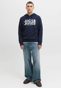 Marineblauer Hoodie mit "JACK & JONES" Grafikdruck, kombiniert mit hellblauen weiten Jeans und braunem Schuhwerk. Schlichtes Design, lockere Passform.