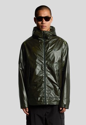 Mand iført mørkegrøn skinnende hættejakke og sorte solbriller, stående mod en lys grå baggrund.