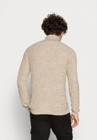 Mann mit lockigem Haar, der einen beigen Strickpullover und schwarze Cargo-Hosen trägt, steht mit dem Rücken zu einem schlichten hellen Hintergrund.