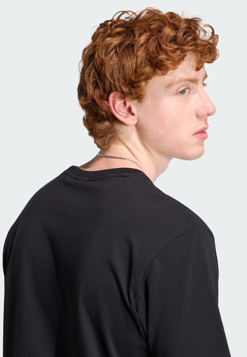 Junge Person mit lockigem roten Haar, die ein schwarzes Shirt und eine silberne Halskette trägt und seitlich vor einem schlichten hellen Hintergrund schaut.
