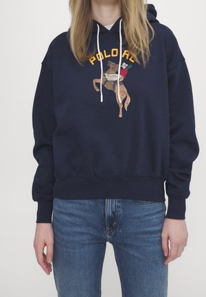 Personne portant un sweat à capuche bleu marine avec un graphisme cheval et cavalier et un texte jaune, associé à un jean bleu, debout devant un fond uni.