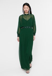 Robe maxi en chiffon vert avec une jupe plissée, des manches longues et une taille et des poignets ornés de sequins, présentée avec un col haut.