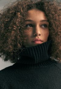 Giovane con capelli ricci che indossa un spesso maglione nero a collo alto, che guarda pensieroso di lato su uno sfondo neutro.