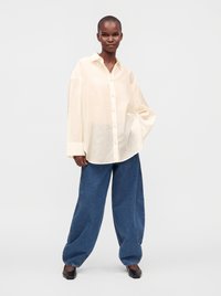 Personne debout, main dans la poche, portant une chemise crème ample boutonnée, un jean large bleu et des chaussures noires pointues sur fond blanc.
