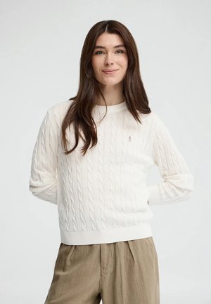 Vrouw met lang bruin haar, gekleed in een witte kabelgebreide trui en beige broek, staand met haar handen achter haar rug.
