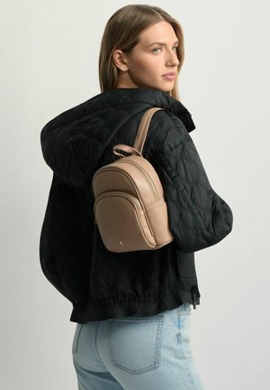 Kobieta ubrana w czarną pikowaną kurtkę i jasnoniebieskie dżinsy, niosąca mały jasnobrązowy skórzany plecak na jednym ramieniu.