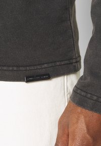 Mörkgrå långärmad tröja med ribbad nederkant och integrerad varumärkesetikett, tillsammans med vita jeans. Slät bomullstextur.