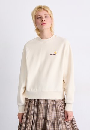 Sweatshirt couleur crème fabriqué en tissu doux, avec un col rond et des poignets côtelés. Un logo brodé sur la poitrine ajoute une touche de détail.