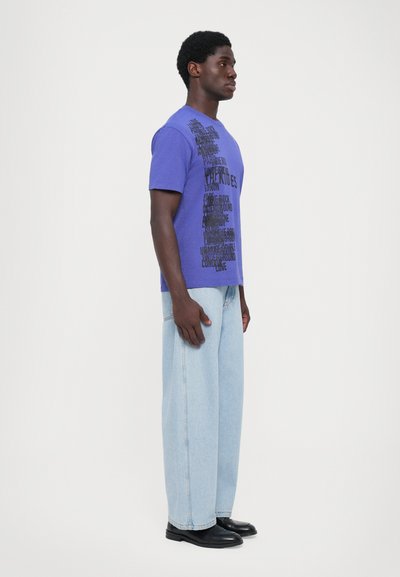 Purple cotton t-shirt with black text design, featuring short sleeves and a crew neckline. Paired with light blue wide-leg jeans and black shoes.