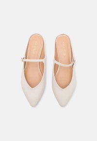 Ett par beiga spetsiga platta skor med tunna ankelremmar och guldspännen på en vit bakgrund.