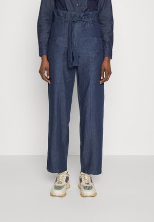 Aikuinen, jolla on päällään tummansiniset korkeavyötäröiset leveälahkeiset housut, joissa on etutaskut ja solmittava vyö, yhdistettynä kermaan ja vihreään paksupohjaisiin lenkkareihin.