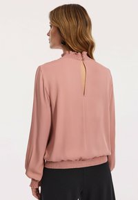 Różowa bluzka z falbaną przy wysokim dekolcie, z wycięciem z tyłu oraz elastycznymi mankietami i dołem. Miękka tkanina o gładkiej teksturze, połączona z czarnymi spodniami.