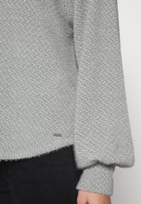 Gray textured sweater featuring a woven pattern, slightly flared sleeves, and a small metallic brand tag near the hem. Black pants visible below.