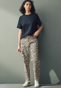 Černé tričko s krátkým rukávem v kombinaci s béžovými a černými pruhovanými kalhotami. Model má na sobě bílé tenisky. Pozadí je jemně zelené.