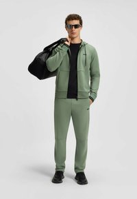 Green athletic tracksuit with a zip-up hoodie and matching pants, accented with black detailing, paired with a black duffle bag and sunglasses.