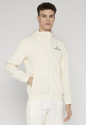 Ung mann iført en lys beige hettejakke med glidelås og logo på brystet, og matchende lyse bukser, stående med den ene hånden i lommen.