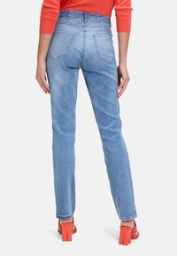 Jeans azzurri a vita alta con taglio dritto, dotati di tasche posteriori e dettagli cuciti, indossati con sandali arancioni a blocchi con tacco.