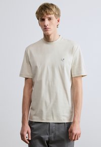 Μπεζ βαμβακερό t-shirt με στρογγυλή λαιμόκοψη, κοντά μανίκια και μικρή λεπτομέρεια λογότυπου στο αριστερό στήθος. Ομαλή υφή, casual εφαρμογή.