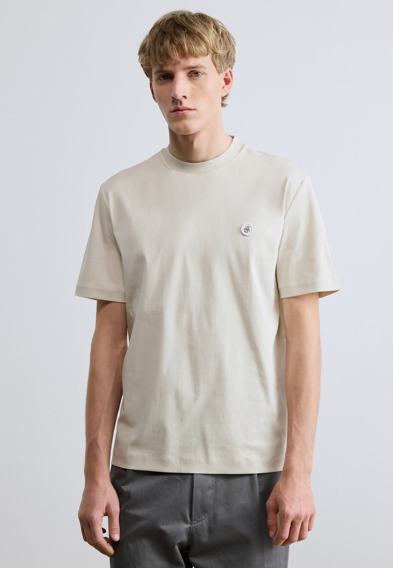 Μπεζ βαμβακερό t-shirt με στρογγυλή λαιμόκοψη, κοντά μανίκια και μικρή λεπτομέρεια λογότυπου στο αριστερό στήθος. Ομαλή υφή, casual εφαρμογή.