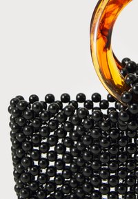 Črna torbica iz perlic z elegantno obliko, ki se ponaša s prozornim jantarnim krožnim ročajem. Gladka tekstura z bleščečo površino.