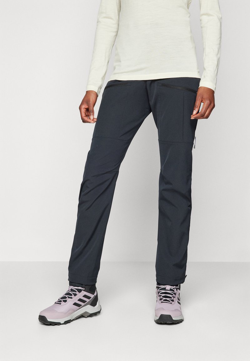 Pantalons noirs d'extérieur avec de multiples poches zippées, fabriqués à partir d'un matériau léger, associés à des chaussures de randonnée violettes. Présente un design épuré.
