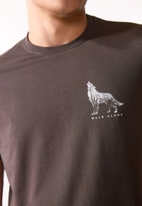 Donkerbruine katoenen T-shirt met een witte wolfafbeelding met boomdetails en de tekst "WALK ALONE" onder het ontwerp.