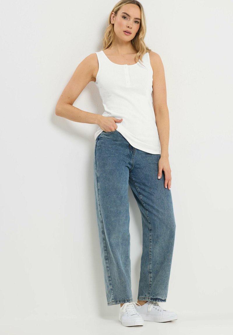 Kvinde med blondt hår iført en hvid ærmeløs top, blå jeans med høj talje og hvide sneakers, stående mod en hvid væg.