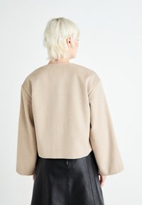 Giacca cropped in misto lana beige con maniche larghe e scollo rotondo, abbinata a una gonna in pelle nera con dettagli di cucitura visibili.