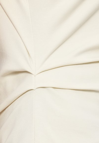 Ύφασμα σε κρεμ χρώμα με συγκεντρωμένες, στριφτές λεπτομέρειες στο κέντρο, που αναδεικνύουν λεία υφή και διακριτική γυαλάδα. Είναι ορατές οι ραφές.