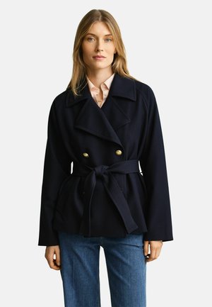 Frau mit blondem Haar, die einen marineblauen, doppelt geknöpften Mantel mit Gürtel über einem hellrosa Hemd und blauen Jeans vor einem einfarbigen Hintergrund trägt.