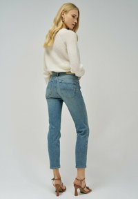 Jeans de mezclilla azul de talle alto con pierna ajustada, que presentan bolsillos traseros y un ligero desvanecimiento. Combinados con un suéter de punto crema y tacones marrones.