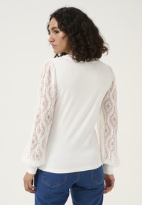 White long-sleeve shirt with lace puff sleeves, a fitted bodice, and a round neckline, paired with blue denim jeans.