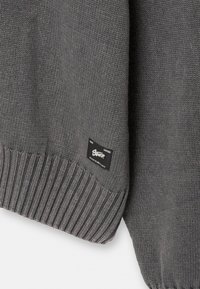 Manche de pull gris tricoté avec un bord-côte et une petite étiquette en tissu noire comportant un texte blanc et un logo près du bord.